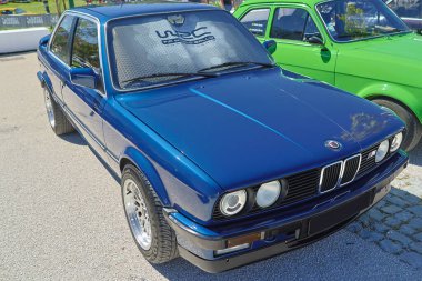 Parlak mavi metalik BMW 3 serisi E30 güneşli bir günde asfalt bir yüzeye park edilmiş.