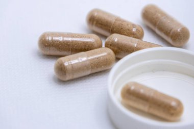 Beyaz bir şişeden beyaz bir arka plana birkaç tane kahverengi bitkisel takviye hapı dökülüyor.