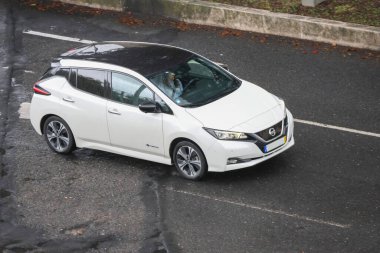 Beyaz Nissan yapraklı, direksiyonda bir şoförle yolda giden elektrikli araba.