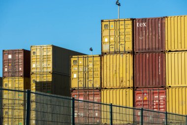 Metal shipping containers stacked high behind a chain link fence at a port terminal, ready for global transport clipart