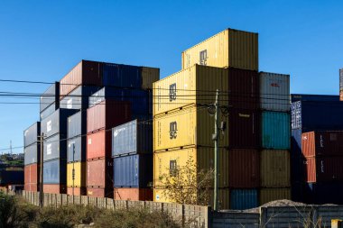 Küresel ticaret ve lojistiği temsil eden açık mavi gökyüzünün altında beton bir duvarın arkasında renkli konteynırlar yığılıyor.
