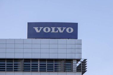 Volvo logosu modern bir binanın tepesinde berrak mavi bir gökyüzüne karşı, markanın varlığını sembolize ediyor.