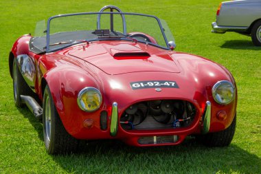 Klasik kırmızı ac kobra 427 roadster yeşil çimen tarlasına park etmiş, zamansız tasarımını ve güçlü motorunu sergiliyor.