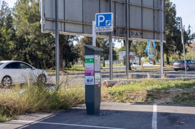 Otopark parkında park taksimetresi ödemeyi bekliyor. Arabalar geçiyordu ve reklam panoları arka plandaydı. Şehir hareketliliğini ve park yönetimini simgeliyordu.