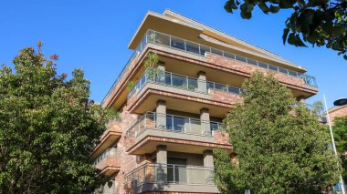 Mavi gökyüzünün altında balkonları ve ağaçları olan modern tuğla bina.