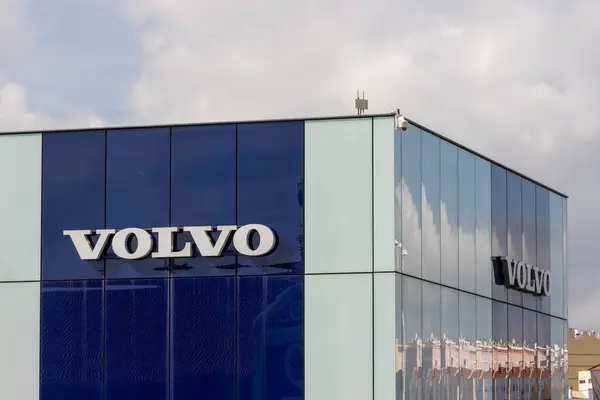 Volvo logosu, yukarıdaki bulutlu gökyüzünü yansıtan modern cam bir binada sergilenmektedir.