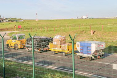Havaalanı bagaj arabaları Lisbon Humberto Delgado Uluslararası Havaalanı 'ndaki pistte bagajları ve kargoları çekiyor. Arka planda uçak iniyor.