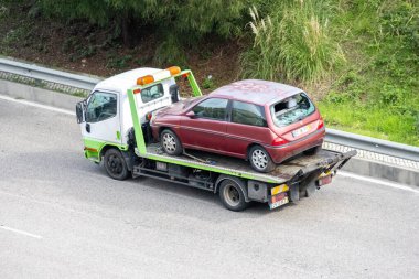 Çekici, otoyolda kırık arka camlı hasarlı kırmızı bir araba taşıyor.