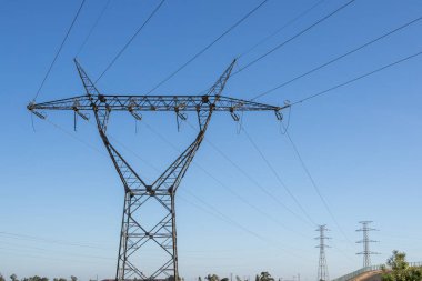 Metal elektrik pilonu ve elektrik hatları açık mavi gökyüzüne hakim, güç dağılımını ve altyapıyı sembolize ediyor
