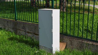 Gri metal elektrikli dolap yeşil bir çitin yanında duruyor. Çimenli bir alanda, muhtemelen bir halk parkında.