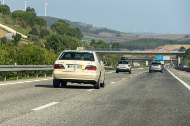 Arka planda rüzgar türbinleri ve diğer arabalarla otoyolda giden bej bir taksi.