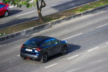 Dark blue citroen c4 driving on the asphalt road in the city with another car and trees in the background clipart