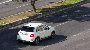 Modern beyaz Fiat 500x araba güneşli bir günde bir ağacın yanında asfalt yolda gidiyor.