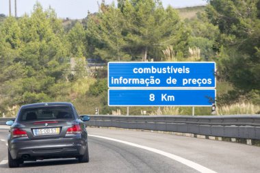 Otoyolda giden koyu gri araba Portekiz 'de 8 kilometre ilerideki benzin fiyatına yaklaşıyor.