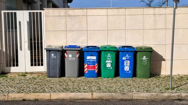 Kaldırımda, fayanslı bir duvarın yanında duran farklı renkli atık kutuları, organize geri dönüşüm ve atık yönetimini destekliyor.