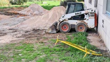 Vaşak kızak dümen yükleyicisi bir kum yığınının yanına park etmiş ve inşaat alanında sarı bir bariyer düşmüş.