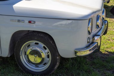 Çimenli bir alana park etmiş beyaz bir Renault 8 'in ön tekerleğini, tamponunu ve farlarını vurguluyor.