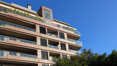 Balkonların, bitkilerin ve panjurların bulunduğu modern tuğla bina, açık mavi gökyüzüne karşı kurulmuş.