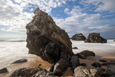 Sintra, Portekiz 'deki Adraga Sahili' nin köpüklü sulardan çıkan taşlı uzun pozlu fotoğrafı.