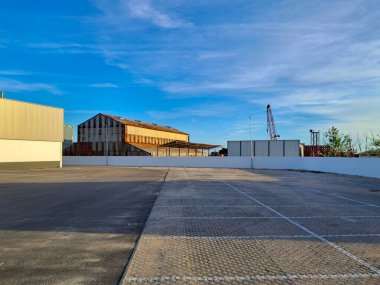 Endüstriyel alanda boş bir otopark, çeşitli binalar ve arka planda açık mavi bir gökyüzünün altında bir vinç var.