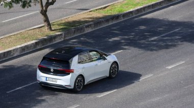 Modern beyaz elektrikli Volkswagen id.3 şehir ortamında asfalt bir yolda sürerken sürdürülebilir ulaşım sergiliyor.