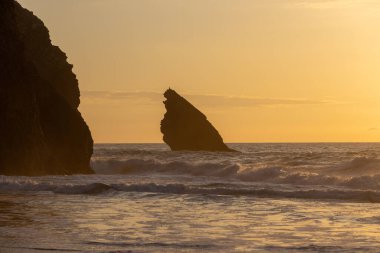 Gün batımında Adraga plajına vuran dalgalar, ikonik pedra da agulha kaya oluşumuyla altın gökyüzüne çarpıyor.
