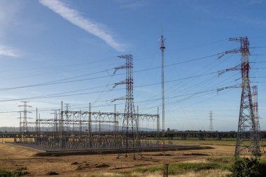 Elektrik santrali, açık mavi gökyüzü altında yüksek voltaj kuleleri ve elektrik hatlarından oluşan bir ağ üzerinden enerji dağıtıyor.