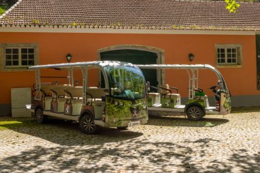 Tapada Nacional de Mafra, Portekiz 'de turuncu bir binanın önüne park edilmiş iki elektrikli servis otobüsü sürdürülebilir ulaşım imkanı sunuyor.