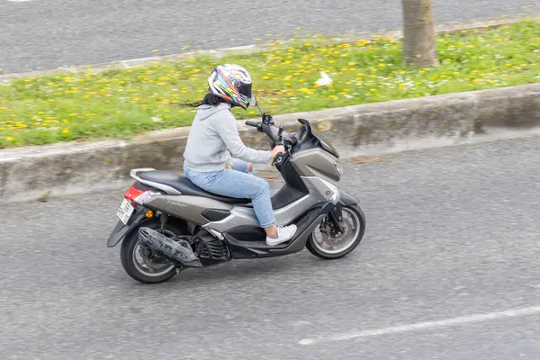 Genç bir kadın asfalt bir yolda gri bir Yamaha xmax scooter kullanıyor çimenli bir kaldırımın yanında