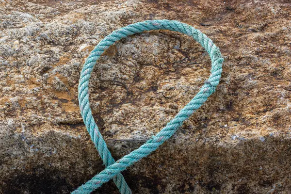 Bir turkuaz halatı kapayarak sert bir kaya yüzeyinde çember oluşturarak, denizcilik ya da demirleme temasını işaret eder.