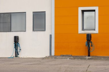 Her iki şarj istasyonu yeşil taşımayı sembolize eden canlı turuncu ve beyaz bir binaya karşı hazır bekliyor.