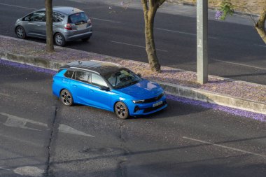 Asfalt yolda yürüyen mavi bir opel astra spor turnuvacısı ve kaldırımda düşmüş jakaranda çiçekleri.