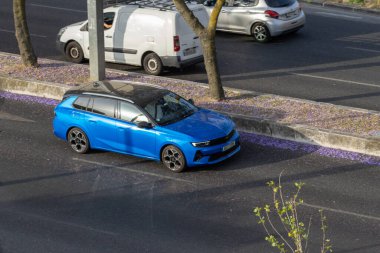 Mavi bir opel astra spor turnuvacısının kaldırımda düşmüş bir jakaranda çiçeğiyle araba sürüşünün hava görüntüsü.