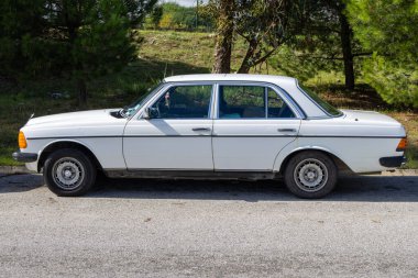 Klasik beyaz Mercedes Benz w123 sedan. Asfaltta, ağaçların yanında park edilmiş.