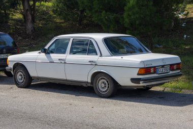 Klasik beyaz Mercedes Benz W123 yol kenarına park etmiş, klasik tasarımını sergiliyor.