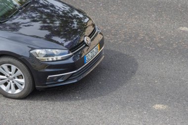 Siyah bir Volkswagen golfün ön tarafı asfalt yolda park edilmiş, kaputuna ağaçlar yansıyor.