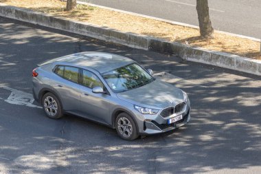 Parlak gümüş bmw, asfalt bir yolda park edilmiş. Yakındaki ağaçların arasından süzülen benekli güneş ışığının altında bir gölge bırakıyor.