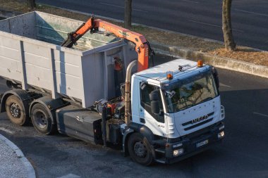 Çöp kamyonu, büyük metal bir konteynırı kaldırıyor ve atık toplama operasyonu yapıyor.