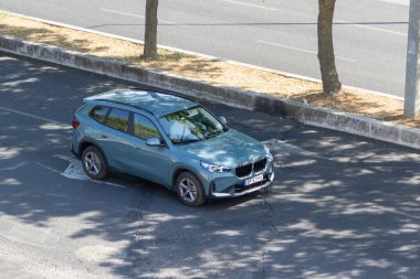 Açık mavi bmw x1 SUv ağaçlık bir yolun kenarına park etmiş, güneşli bir günde asfaltta gölgeler bırakıyor.