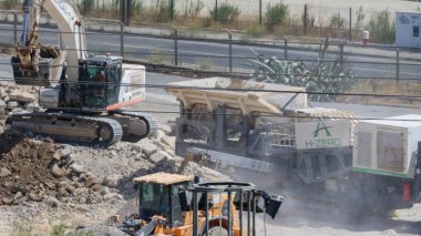 Kazıcı ezilmiş taşları seyyar öğütücüye yüklüyor, yol inşaatı sırasında diğer ağır makinelerle birlikte çalışıyor.