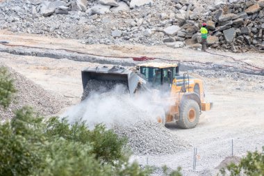 Sarı tekerlek yükleyicisi arka planda su püskürten bir inşaat işçisiyle taş ocağına çakıl boşaltıyor.