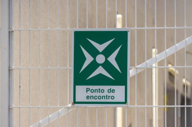 Green assembly point sign indicating safe gathering location during emergencies, attached to a metal fence, ensuring clear visibility and guidance