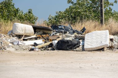 Pile of garbage polluting the environment including mattress, plastic, and furniture near trees