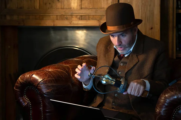 A man in a hat and a Victorian-era costume with an antique lamp in his hand looks at the laptop in a retro style. The theme is modern technology and the last century.