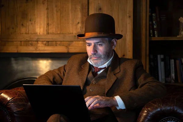 A man in a hat and a Victorian costume works with a laptop in a salon in retro style. The theme is modern technology and past century.