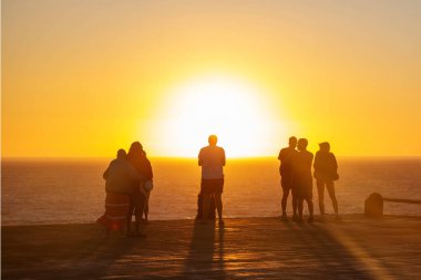 Parlak günbatımına karşı iskelede duran insanların siluetleri. Gölgeleri gün batımında okyanus ufkunda aydınlanan bir grup insan..