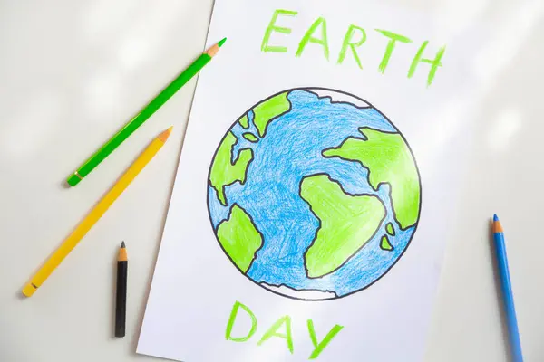 A white sheet of paper depicting a globe painted with colored pencils and the inscription Earth Day is on the table lit by daylight.