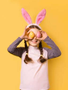 Paskalya tavşanı kulakları ve sarı arka planda renkli Paskalya yumurtaları olan neşeli bir kız. Mutlu Paskalyalar!