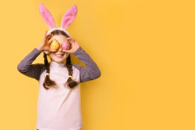 Paskalya tavşanı kulakları ve sarı arka planda renkli Paskalya yumurtaları olan neşeli bir kız. Mutlu Paskalyalar! Boşluğu kopyala.