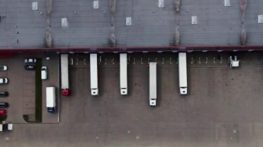Semi truck with cargo trailer is travelling along a parking lot of a warehouse in the logistics park. A lot of semi-trailers trucks stands at warehouses ramps for load/unload goods. Aerial view